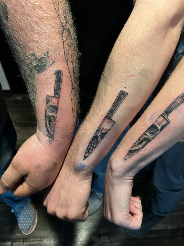 Matching black and grey realism chef-knife tattoos with skulls inside the blades on three forearms