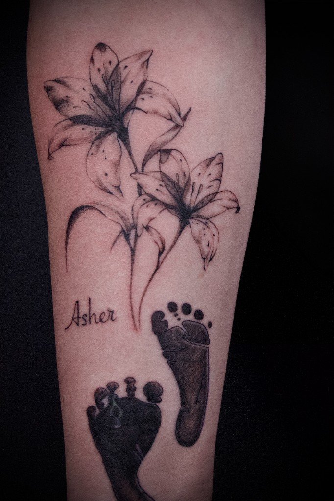 Fine-line lilies above two inked newborn footprints and the name Asher, on a forearm