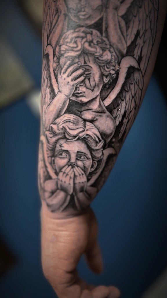 Black and grey realism cherub angels tattoo covering a forearm, one covering its eyes, one its mouth