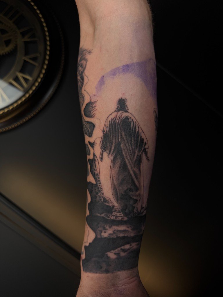 Black and grey tattoo of a robed figure seen from behind, standing on rocks under a pale sky, forearm